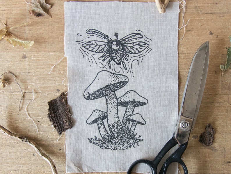 May include: A rectangular piece of light beige fabric featuring a black ink illustration of a cluster of mushrooms with a beetle flying above them. The design uses stippling and line work to create texture. A pair of metal scissors is positioned nearby.