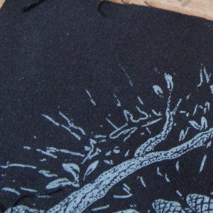 May include: A black fabric patch featuring a screen-printed design in light blue ink. The graphic depicts a stylized tree with textured branches and leaves, rendered in a distressed, hand-drawn aesthetic with visible fabric weave texture.