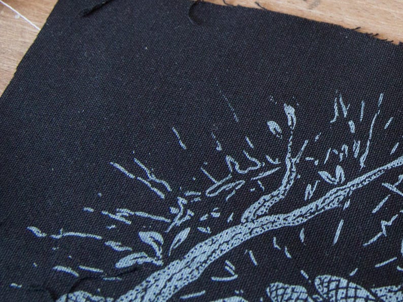 May include: A black fabric patch featuring a screen-printed design in light blue ink. The graphic depicts a stylized tree with textured branches and leaves, rendered in a distressed, hand-drawn aesthetic with visible fabric weave texture.