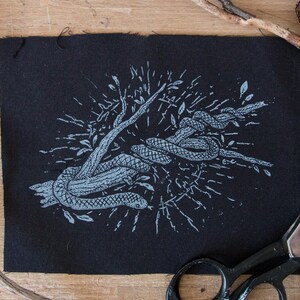 May include: A rectangular black fabric patch featuring a light blue screen-printed illustration of a snake coiled around a tree branch. The design includes radiating lines and small leaf motifs surrounding the central snake and branch imagery.