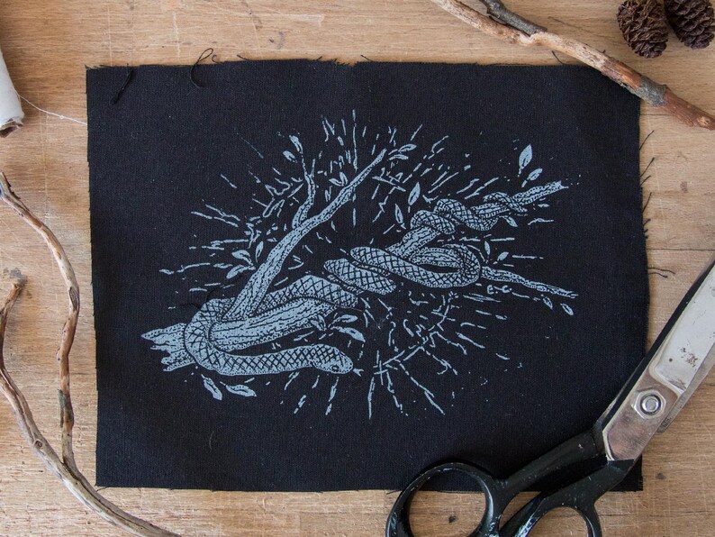 May include: A rectangular black fabric patch featuring a light blue screen-printed illustration of a snake coiled around a tree branch. The design includes radiating lines and small leaf motifs surrounding the central snake and branch imagery.