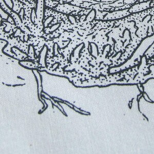 May include: A close-up view of a light-colored fabric featuring a black ink illustration. The design displays a textured, organic shape with intricate line work, stippling, and small root-like extensions reaching downward from the main form.