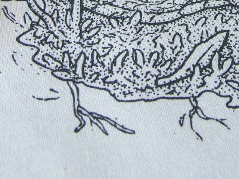 May include: A close-up view of a light-colored fabric featuring a black ink illustration. The design displays a textured, organic shape with intricate line work, stippling, and small root-like extensions reaching downward from the main form.
