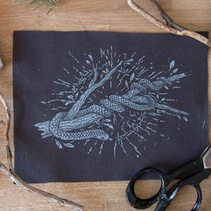 May include: A rectangular dark gray fabric patch featuring a light blue screen-printed illustration of a snake coiled around a tree branch. The design includes radiating lines and small leaves surrounding the central snake and branch motif.