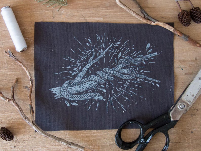 May include: A rectangular dark gray fabric patch featuring a light blue screen-printed illustration of a snake coiled around a tree branch. The design includes radiating lines and small leaves surrounding the central snake and branch motif.