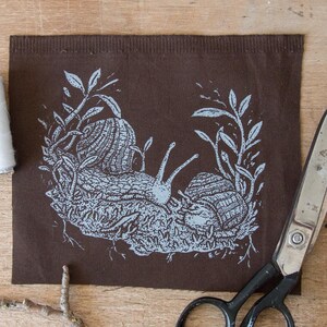 May include: A rectangular piece of dark brown fabric featuring a light blue screen-printed illustration of two snails crawling among foliage and roots. The fabric is positioned next to a pair of metal scissors and two spools of sewing thread.