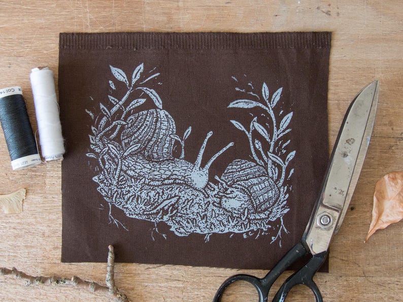 May include: A rectangular piece of dark brown fabric featuring a light blue screen-printed illustration of two snails crawling among foliage and roots. The fabric is positioned next to a pair of metal scissors and two spools of sewing thread.