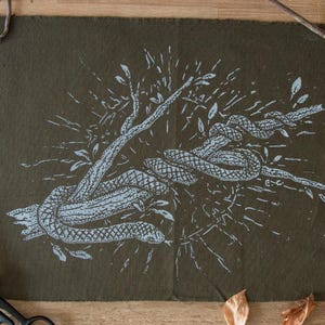 May include: An olive green textile print featuring a detailed illustration of a snake coiled around a tree branch. The snake and branch are rendered in a light gray color. The artwork is surrounded by twigs, thread spools, and scissors.