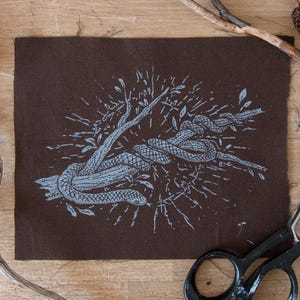 May include: A rectangular piece of dark brown fabric featuring a light blue screen-printed illustration of a snake coiled around a tree branch. The design includes radiating lines and small leaf motifs surrounding the central snake and branch graphic.