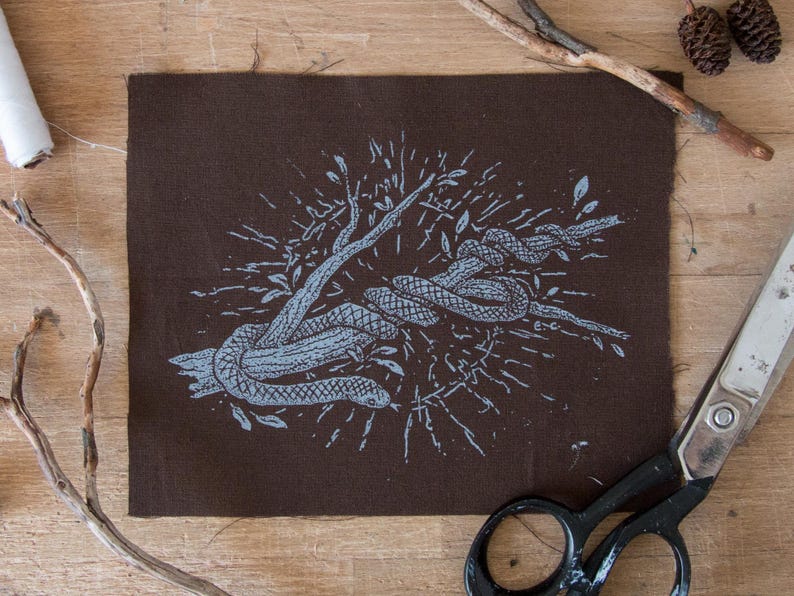 May include: A rectangular piece of dark brown fabric featuring a light blue screen-printed illustration of a snake coiled around a tree branch. The design includes radiating lines and small leaf motifs surrounding the central snake and branch graphic.
