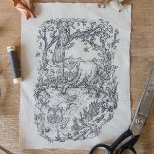May include: A black and white linocut print on fabric showing a raccoon in a forest scene with a pond. The print includes a raccoon in the water and another on the bank. A spool of thread and scissors are next to the print.