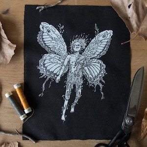 Può includere: Una toppa rettangolare in tessuto nero con un'illustrazione serigrafata bianca di una figura con grandi e dettagliate ali di farfalla. Il disegno mostra la figura in una posa fluttuante con un intricato lavoro di linee e le iniziali E.C.