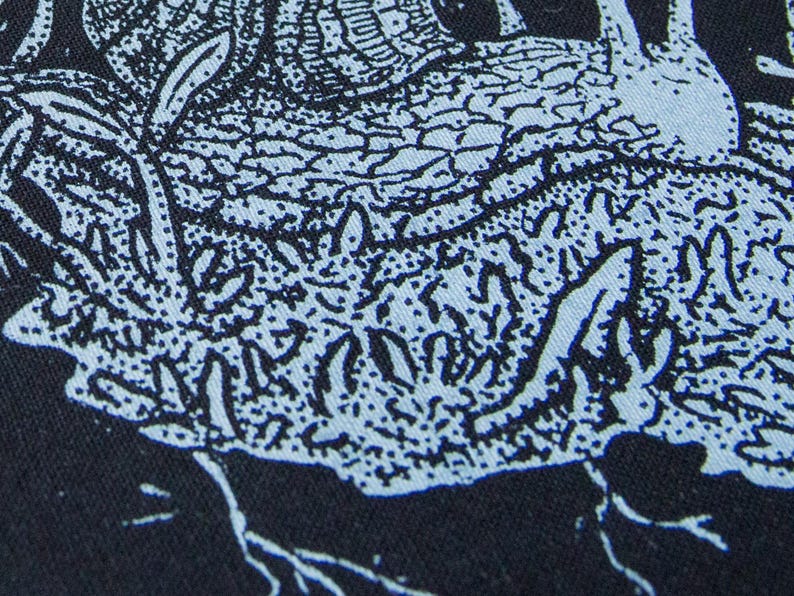 May include: A black fabric featuring a light blue screen-printed graphic of a snail with a textured shell. The design includes intricate line work depicting the snail moving across a patch of ground with small plants and roots visible below.