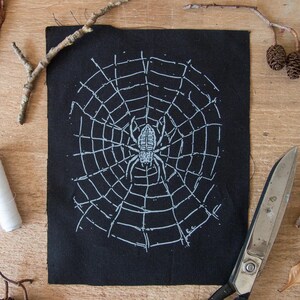Peut inclure: Un écusson rectangulaire en tissu noir présentant un motif sérigraphié blanc représentant une araignée au centre d'une toile circulaire. L'araignée a huit pattes et un corps détaillé. Les initiales E.C. sont imprimées en blanc en bas à droite.