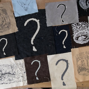 May include: A collection of rectangular fabric patches featuring various screen-printed designs, including question marks in different colors, a bear with the text DAMN HUMANS, a snail, a mole in a nest, and a person sitting with the text TAKE ROOT.