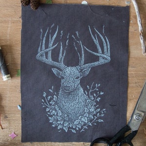 Puede incluir: Una tela gris oscuro con una ilustración azul claro de la cabeza de un ciervo, rodeada de elementos florales. El ciervo tiene grandes astas. La imagen está sobre una superficie de madera con tijeras, carretes de hilo y hojas.