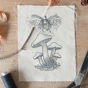 May include: A rectangular piece of off-white fabric featuring a black ink illustration of a cluster of mushrooms and a flying beetle above them. The fabric has raw, frayed edges and includes the initials E.C. printed near the bottom right corner.