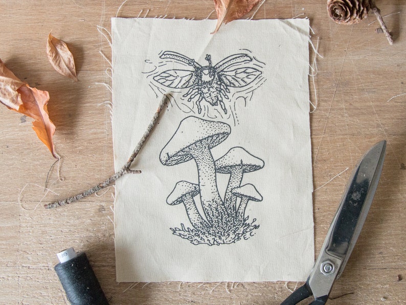May include: A rectangular piece of off-white fabric featuring a black ink illustration of a cluster of mushrooms and a flying beetle above them. The fabric has raw, frayed edges and includes the initials E.C. printed near the bottom right corner.