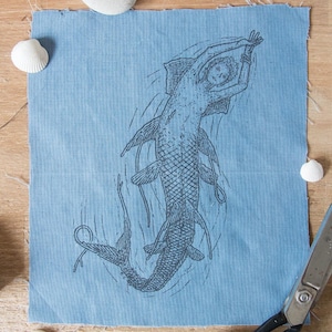 Könnte beinhalten: Ein quadratisches Stück hellblauer Stoff mit einer detaillierten schwarzen Tuschezeichnung einer Meerjungfrau mit Fischschwanz und Flossen. Das Motiv zeigt die Figur beim Schwimmen mit erhobenen Armen, umgeben von feinen Linien, die Wellen darstellen.