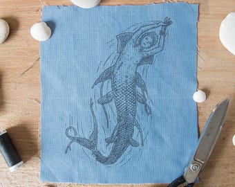 May include: A square piece of light blue fabric featuring a detailed black ink illustration of a mermaid with a fish-like tail and fins. The design shows the figure swimming with arms raised above the head, surrounded by subtle water ripple lines.