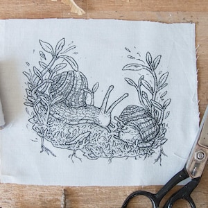 May include: A rectangular piece of off-white fabric featuring a black ink illustration of two snails surrounded by foliage and moss. The design includes the initials E.C. in the bottom right corner. Two spools of thread and metal scissors are present.