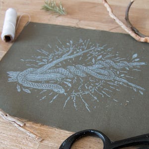 May include: A square piece of olive green fabric featuring a light blue screen-printed illustration of a snake coiled around a branch. The design includes radiating lines and small leaves surrounding the central snake and branch motif.