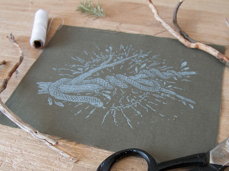May include: A square piece of olive green fabric featuring a light blue screen-printed illustration of a snake coiled around a branch. The design includes radiating lines and small leaves surrounding the central snake and branch motif.