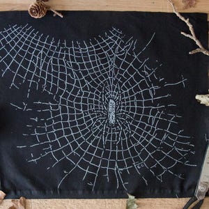 Könnte beinhalten: Ein rechteckiges Stück schwarzer Stoff mit einer weißen, gestickten Spinne in der Mitte eines detaillierten Netzes. Das Design bedeckt den Großteil der Stoffoberfläche und zeigt komplizierte Linien, die das Netz und den Körper der Spinne bilden.