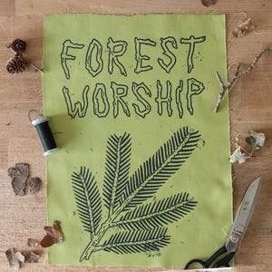 Könnte beinhalten: Ein grüner Stoff mit den schwarzen Worten "FOREST WORSHIP" und einem schwarzen Farn-Design. Das Bild enthält eine Schere, Faden, Tannenzapfen und Zweige auf einer Holzoberfläche.