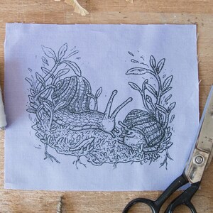 May include: A rectangular piece of light purple fabric featuring a black ink illustration of two snails resting on a patch of moss surrounded by leafy stems. The fabric is positioned next to a spool of black thread, white thread, and metal scissors.