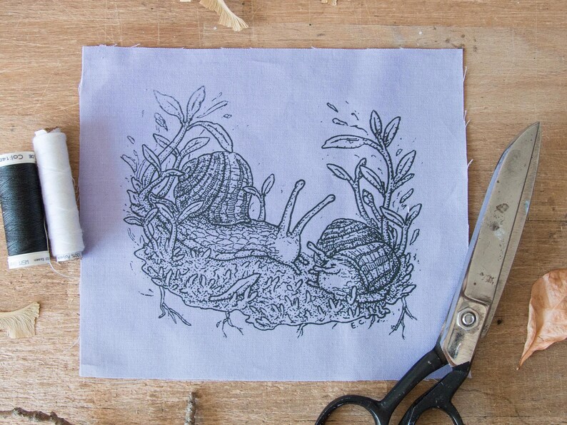 May include: A rectangular piece of light purple fabric featuring a black ink illustration of two snails resting on a patch of moss surrounded by leafy stems. The fabric is positioned next to a spool of black thread, white thread, and metal scissors.