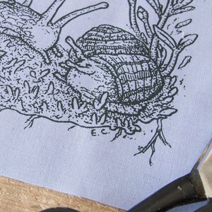 May include: A light blue fabric panel featuring a black ink illustration of a snail with a textured shell crawling on mossy ground. The drawing includes small plant details and the initials E.C. near the bottom. Metal scissors are visible.