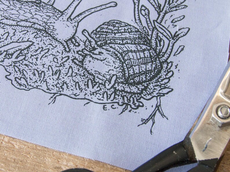 May include: A light blue fabric panel featuring a black ink illustration of a snail with a textured shell crawling on mossy ground. The drawing includes small plant details and the initials E.C. near the bottom. Metal scissors are visible.