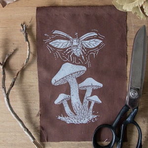 May include: A rectangular brown fabric patch featuring a white screen-printed design of a cluster of three mushrooms at the bottom and a winged insect at the top. The initials E.C. are printed in white near the base of the mushroom cluster.