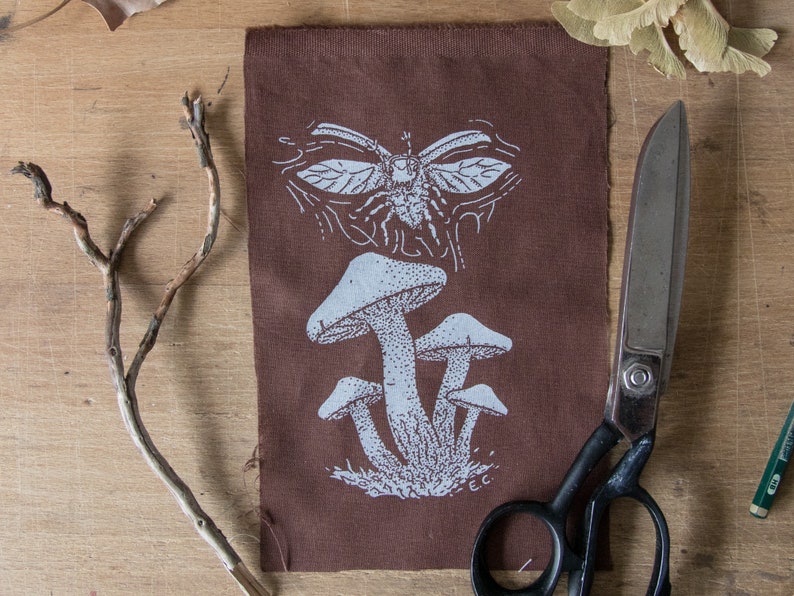 May include: A rectangular brown fabric patch featuring a white screen-printed design of a cluster of three mushrooms at the bottom and a winged insect at the top. The initials E.C. are printed in white near the base of the mushroom cluster.