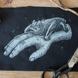 May include: A rectangular black fabric patch featuring a white screen-printed illustration of a bat resting on top of a human hand. The design uses a stippled dot-work style and includes small star-like accents surrounding the central figures.