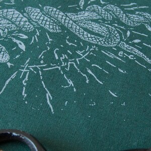 May include: A dark green fabric featuring a white screen-printed illustration of intertwined snakes and foliage. The design includes fine line work and a small E.C. signature mark. A portion of a black circular object is visible at the bottom edge.
