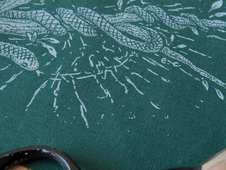 May include: A dark green fabric featuring a white screen-printed illustration of intertwined snakes and foliage. The design includes fine line work and a small E.C. signature mark. A portion of a black circular object is visible at the bottom edge.