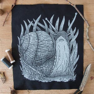 Könnte beinhalten: Ein schwarzer Stoffaufnäher mit einer detaillierten, weiß siebgedruckten Illustration einer Schnecke, die durch hohes Gras kriecht. Die Schnecke hat ein strukturiertes Gehäuse und sichtbare Augenstiele, dargestellt in einem punktierten Stil.