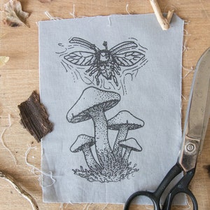 May include: A rectangular piece of light gray fabric featuring a black ink illustration of a cluster of mushrooms and a flying beetle above them. The fabric has raw, frayed edges and is positioned next to a pair of black metal scissors.