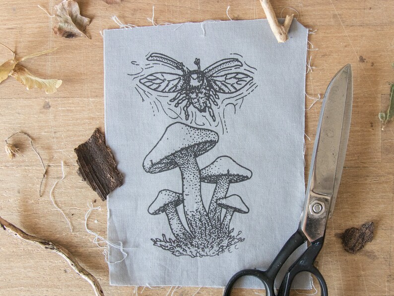 May include: A rectangular piece of light gray fabric featuring a black ink illustration of a cluster of mushrooms and a flying beetle above them. The fabric has raw, frayed edges and is positioned next to a pair of black metal scissors.