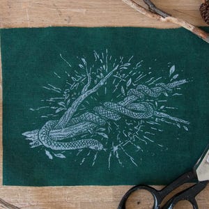 May include: A rectangular piece of dark green fabric featuring a white screen-printed illustration of a snake coiled around a branch. The design includes radiating lines and small leaf details surrounding the central snake and branch motif.