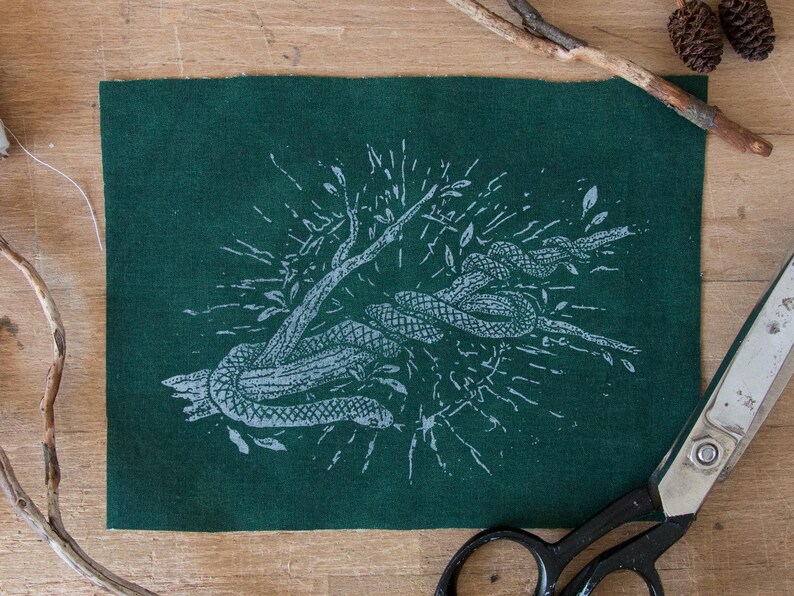 May include: A rectangular piece of dark green fabric featuring a white screen-printed illustration of a snake coiled around a branch. The design includes radiating lines and small leaf details surrounding the central snake and branch motif.