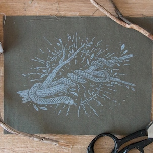 May include: A rectangular piece of olive green fabric featuring a light blue screen-printed graphic of a snake coiled around a tree branch. The design includes radiating lines and small leaves surrounding the central snake and branch illustration.