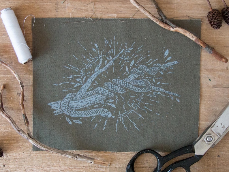 May include: A rectangular piece of olive green fabric featuring a light blue screen-printed graphic of a snake coiled around a tree branch. The design includes radiating lines and small leaves surrounding the central snake and branch illustration.
