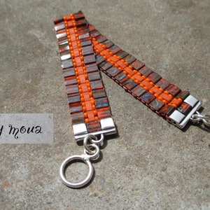 Peut inclure: Un bracelet fait à la main avec un motif orange et marron. Le bracelet est composé de petites perles carrées et de perles orange au centre. Il possède un fermoir argenté. Le texte "by moua" est dans l'image.