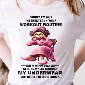 Op de afbeelding: Wit T-shirt met de tekst "SORRY I'M NOT INTERESTED IN YOUR WORKOUT ROUTINE". Het shirt heeft een cartoon illustratie van een vrouw in een roze badjas en slippers. De tekst hieronder luidt "I'M HAPPY JUST GETTING MY LEG THROUGH MY UNDERWEAR WITHOUT FALLING DOWN."