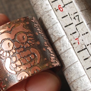 May include: A copper ring with an intricate design of an owl etched into the surface. The ring is being measured with a ruler, showing it is approximately 1 7/8 inches in diameter.