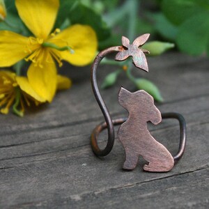 May include: A copper napkin ring with a dog silhouette and leaf detail. The ring has a curled design and a rustic finish. Yellow flowers and green leaves are visible in the background. The ring is made of metal.