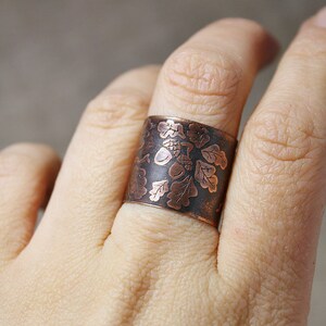 Puede incluir: Un anillo ancho de cobre con un dise&ntilde;o grabado de hojas de roble y bellotas. El anillo se lleva en un dedo.